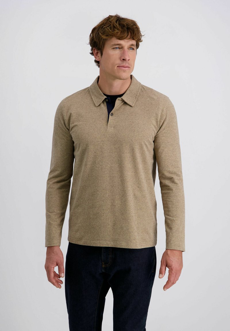 Mann mit lockigem Haar, der ein beiges Langarm-Polohemd und dunkle Jeans trägt und vor einem einfarbigen, hellen Hintergrund steht.