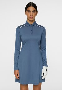 Nainen, jolla on yllään pitkähihainen sininen polo-mekko ja valkoinen golf-käsine vasemmassa kädessään, seisoo yksiväristä taustaa vasten.