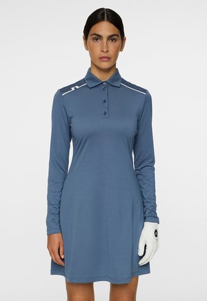 Femme portant une robe polo bleu à manches longues et un gant de golf blanc sur sa main gauche, se tenant contre un fond uni.