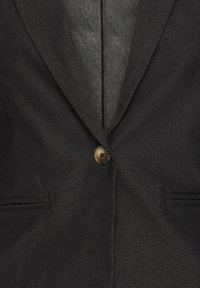 Veste noire avec un seul bouton en laiton, tissu texturé, deux poches latérales et une doublure intérieure lisse. Coupe ajustée avec un col à revers.