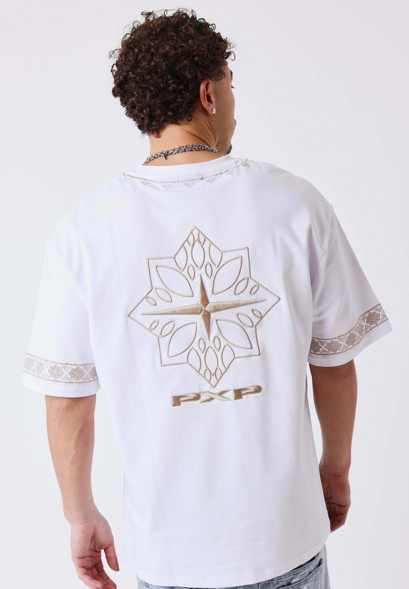 Homme aux cheveux bouclés portant une chemise blanche à manches courtes avec un motif en forme d'étoile géométrique beige et le texte "PXP" au dos, ainsi que des bordures de manches assorties.