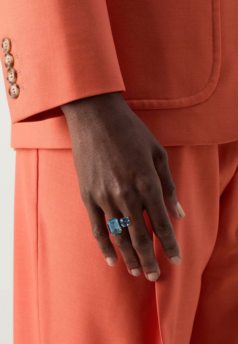 Eine Hand mit zwei Ringen: einem rechteckigen hellblauen Stein und einem runden dunkelblauen Stein, vor einem Hintergrund eines orangenen Anzugs mit Knopfakzenten.