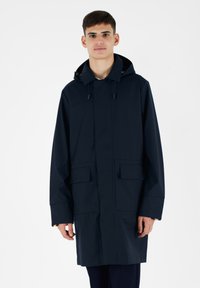 Giacca impermeabile blu navy con cappuccio, zip frontale, due grandi tasche frontali e cordini regolabili. Texture liscia e design moderno.
