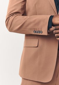 Veste de costume marron à la texture lisse, avec une poche unique et quatre boutons foncés sur le poignet. Coupe cintrée avec des lignes épurées et un design subtil.