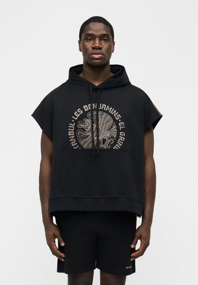 Bărbat purtând hanorac negru fără mâneci cu grafică circulară și text, asortat cu pantaloni scurți negri, stând în fața unui fundal alb simplu.