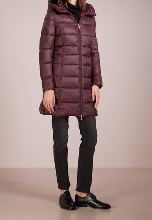 Kobieta w bordowej, puchowej kurtce sięgającej do kolan z kapturem, czarnych dżinsach i czarnych skórzanych butach, stojąca na tle jednolitego tła.