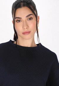 Top blu navy con un motivo a spina di pesce testurizzato, collo rotondo e dettagli di cucitura sottili. Indossato con grandi orecchini leggeri.