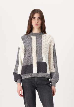 Maglione di maglia spessa con un collo alto, caratterizzato da un design a patchwork in grigio, crema e blu scuro. Ogni sezione presenta texture distintive.