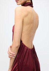 Robe en satin bordeaux avec un dos décolleté, une taille froncée et une fine bretelle autour du cou. Texture lisse avec un éclat subtil.