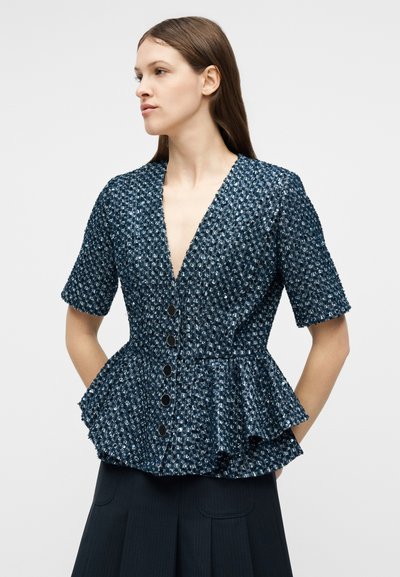 Femme portant un haut à boutons texturé bleu marine à sequins avec ourlet à basque et manches courtes, debout devant un fond uni.