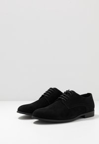 Une paire de chaussures habillées en cuir suédé noir pour hommes, lacées, sur une surface blanche avec un fond gris clair.