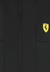 Czarna kurtka z zamkiem błyskawicznym, z żółtym logo w kształcie tarczy przedstawiającym skaczącego konia. Materiał wydaje się gładki o matowym wykończeniu.