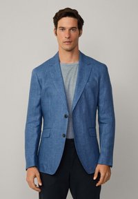 Blazer in lino blu con rever a punta, due bottoni e due tasche frontali. Indossato sopra una camicia grigia a righe, abbinato a pantaloni scuri.