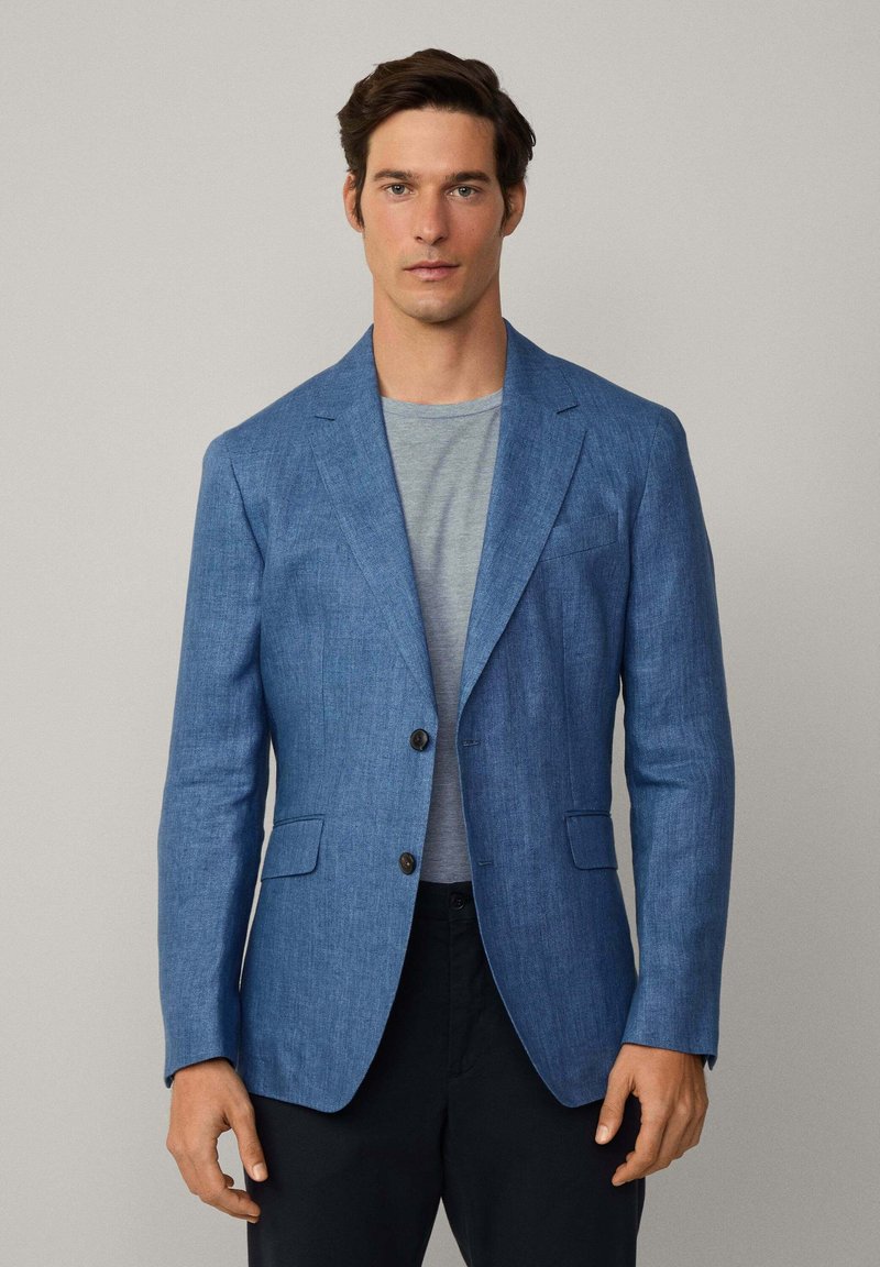 Blazer in lino blu con rever a punta, due bottoni e due tasche frontali. Indossato sopra una camicia grigia a righe, abbinato a pantaloni scuri.