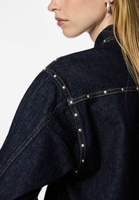 Mørk denimjakke med perlepynt langs skuldersømmene, med kontrastsøm og en strukturert design.