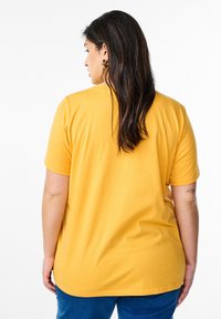 T-shirt à manches courtes, col rond en coton jaune vif. Texture lisse avec une coupe décontractée et un ourlet arrière légèrement plus long.