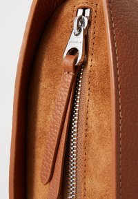 Détail en daim marron avec glissière en cuir, dotée d'un tirette en cuir et d'une fermeture éclair en métal argenté. Le gros plan met en avant les matériaux lisses et texturés.