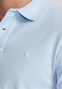 Světle modrá polo košile vyrobená ztexturované bavlny, s klasickým límcem a malým bílým vyšitým logem na levém hrudníku.