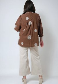 Bruine blouse met witte bloemembroideries, relaxte pasvorm, geplooide halslijn, lange mouwen, gedragen met crèmekleurige wijde broek en platformsandalen.
