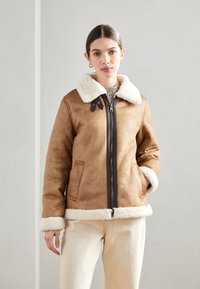 Giacca di shearling marrone con colletto e polsini in pelliccia bianca, dotata di zip frontale e tasche laterali, su uno sfondo neutro.