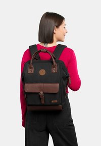 Zaino in tessuto nero con dettagli in pelle marrone, manici superiori e spallacci regolabili. Presenta una patch con logo e un pattina anteriore con una fibbia.
