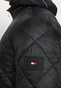 Svart pufferjacka med en diamantquiltad design. Har en hög krage och en logopatch i underkant. Slät, glänsande yta.