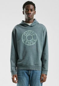 Bluza z kapturem w stonowanym odcieniu teal, z okrągłym graficznym wzorem "Quite Sense" w jasnej zieleni. Miękka textura z prążkowanymi mankietami i dołem.