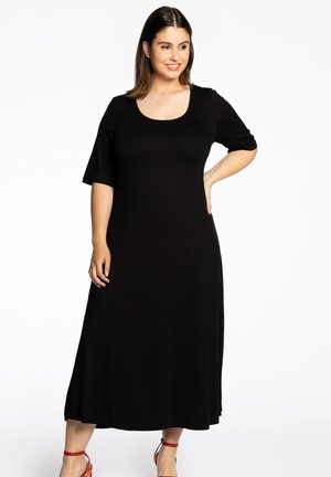 Femme en robe noire midi à manches longueur elbow et talons à cheville rouges, se tenant avec une main sur la hanche, souriant légèrement vers la droite.