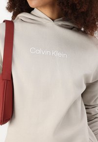 Svetlo béžová mikina s textúrovaným povrchom a kapucňou na šnúrku. Na bielej vyšívanej "Calvin Klein" logo. Viditeľný červený popruh kabelky.