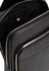 Μαύρο δερμάτινο σακίδιο πλάτης με πολλαπλές θήκες με φερμουάρ, υφή στην επιφάνεια και ταμπέλα με λογότυπο που διαθέτει κόκκινη, λευκή και μπλε λεπτομέρεια.