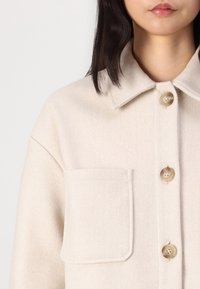 Light beige wool shirt jacket with a pointed collar, three buttons, and a front pocket. Soft texture with a subtle finish.