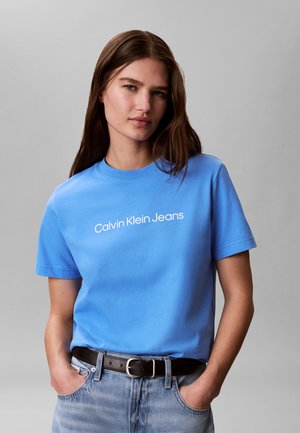 Νεαρή γυναίκα φοράει ένα γαλάζιο t-shirt Calvin Klein Jeans μαζεμένο σε ανοιχτό μπλε τζιν με μαύρη ζώνη, στέκεται με τα χέρια στις τσέπες.