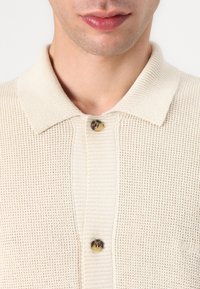 Cream-colored knitted polo shirt with a textured pattern, featuring a button closure and a ribbed collar.