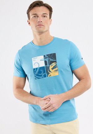 Homme portant un T-shirt bleu clair avec un motif d'étoile de mer, de tentacule de pieuvre et de crabe, debout devant un fond blanc uni.