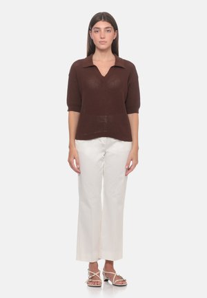 Donna in piedi che indossa un top marrone in maglia a maniche corte con colletto, pantaloni bianchi a gamba larga e sandali bianchi con cinturini su uno sfondo bianco semplice.