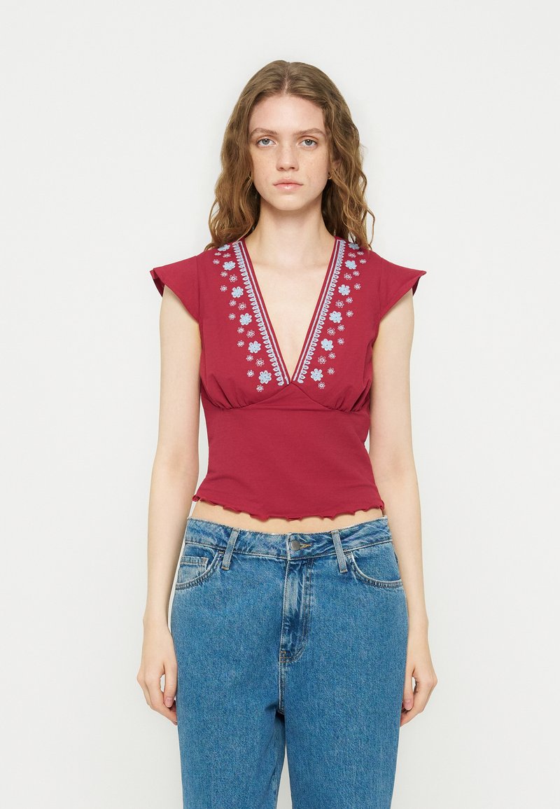 Νεαρή γυναίκα με κόκκινο αμάνικο τοπ με μπλε κεντήματα λουλουδιών και βαθύ V-λαιμόκοψη, συνδυασμένο με τζιν με ψηλή μέση.