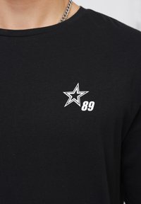 Černé bavlněné tričko s kulatým výstřihem, na kterém je na levé části hrudníku bílé grafické ztvárnění hvězdy a číslo "89".