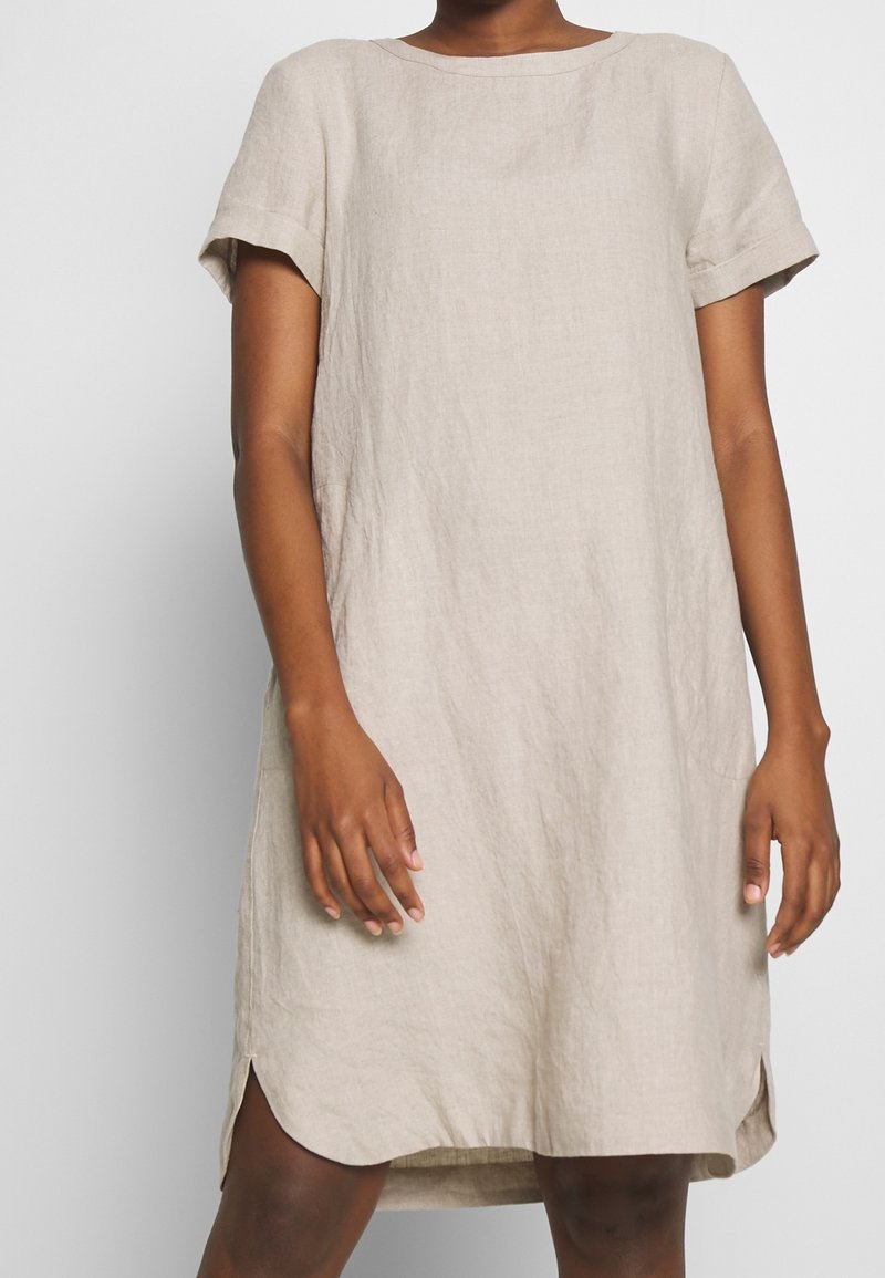 Personne portant une robe en lin beige à manches courtes, coupe ample et ourlet arrondi, debout devant un fond uni clair.
