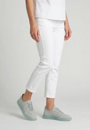 Osoba nosící bílé krátké kalhoty a světle šedé tenisky s průhlednými podrážkami, stojící na jednoduchém světlém pozadí.