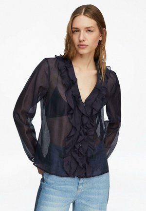 Młoda kobieta ubrana w prześwitującą czarną bluzkę z falbanami z przodu i jasnoniebieskie dżinsy, pozująca z rękami za plecami na białym tle.