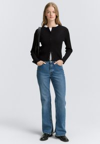 Schwarze gerippte Strickjacke mit Knöpfen, kombiniert mit hellblauen Schlagjeans. Das Model trägt ein weißes Oberteil und schwarze Stiefel und hält eine schwarze Tote-Tasche.