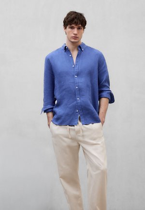 Junger Mann trägt ein blaues, langärmeliges Hemd mit hochgekrempelten Manschetten und beigen, mit Kordelzug versehenen Hosen, steht mit Händen in den Taschen.