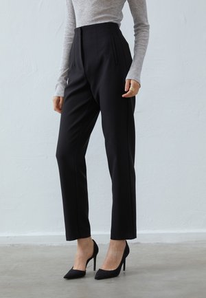 Personne portant un pantalon noir taille haute taillé sur mesure, un haut gris à manches longues, des talons aiguilles noirs pointus, debout contre un mur clair uni.