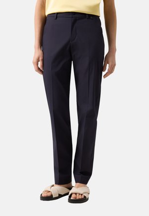 Osoba ubrana w granatowe, dopasowane spodnie i beżowe sandały z krzyżującymi się paskami, widoczna od talii do stóp na jednolitym tle.