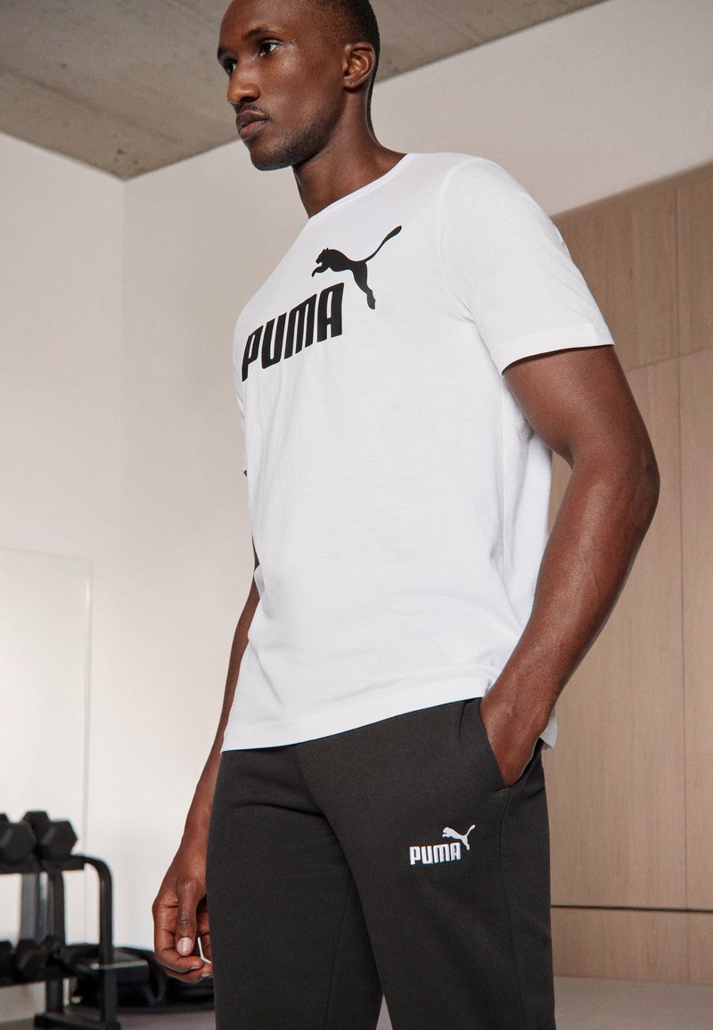 Witte sportieve t-shirt met zwart "PUMA" logo en embleem. Gecombineerd met donkergrijze joggingbroek met een klein logo. Eenvoudig, sportief ontwerp.