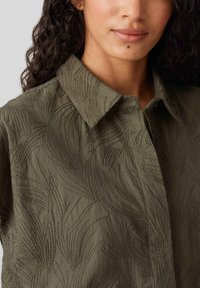 Olijfgroene overhemd met kraag heeft een gestructureerde stof met een bladerenpatroon. Het ontwerp heeft zichtbare stiksels en een relaxte pasvorm.