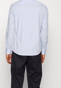 Chemise bleu clair à manches longues avec une texture lisse, fermeture par boutons et ourlet arrondi, associée à un pantalon sombre. Vue de dos visible.