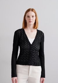 Cardigan noir à manches longues avec un motif en chevron, fermeture à boutons sur le devant et tissu texturé. Porté sur un top blanc et associé à un pantalon crème.