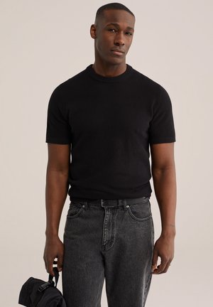 Mand iført en sort strikket kortærmet top og falmede sorte jeans, der holder en sort rygsæk i stropperne mod en ensfarvet baggrund.