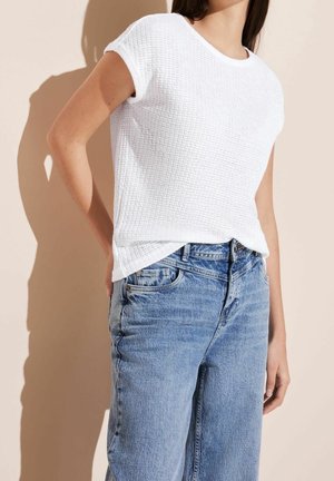 Kobieta w białej, teksturowanej bluzce z krótkim rękawem i jasnoniebieskich, wysokich dżinsach na beżowym tle.